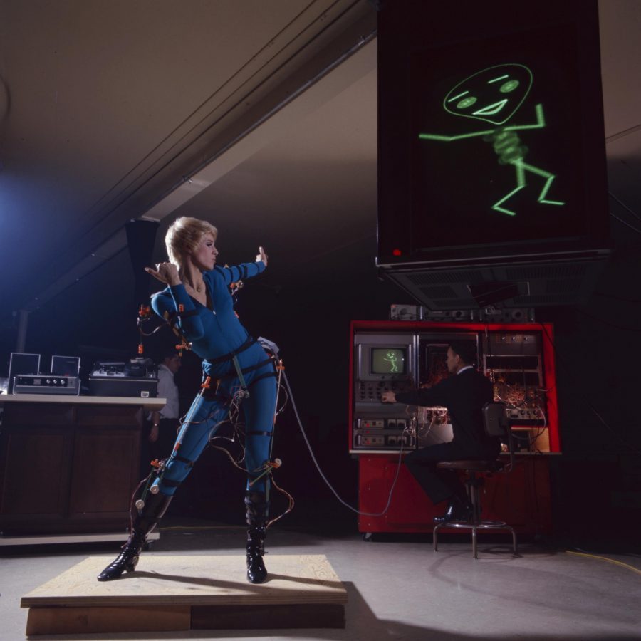 A performer wearing body-mounted sensors controls ANIMAC; a stick figure on the screen above follows her pose